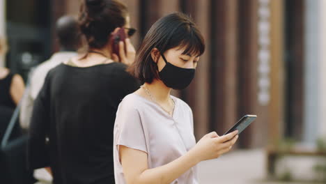 Asian-Woman-in-Mask-Standing-on-Street-and-Using-Smartphone